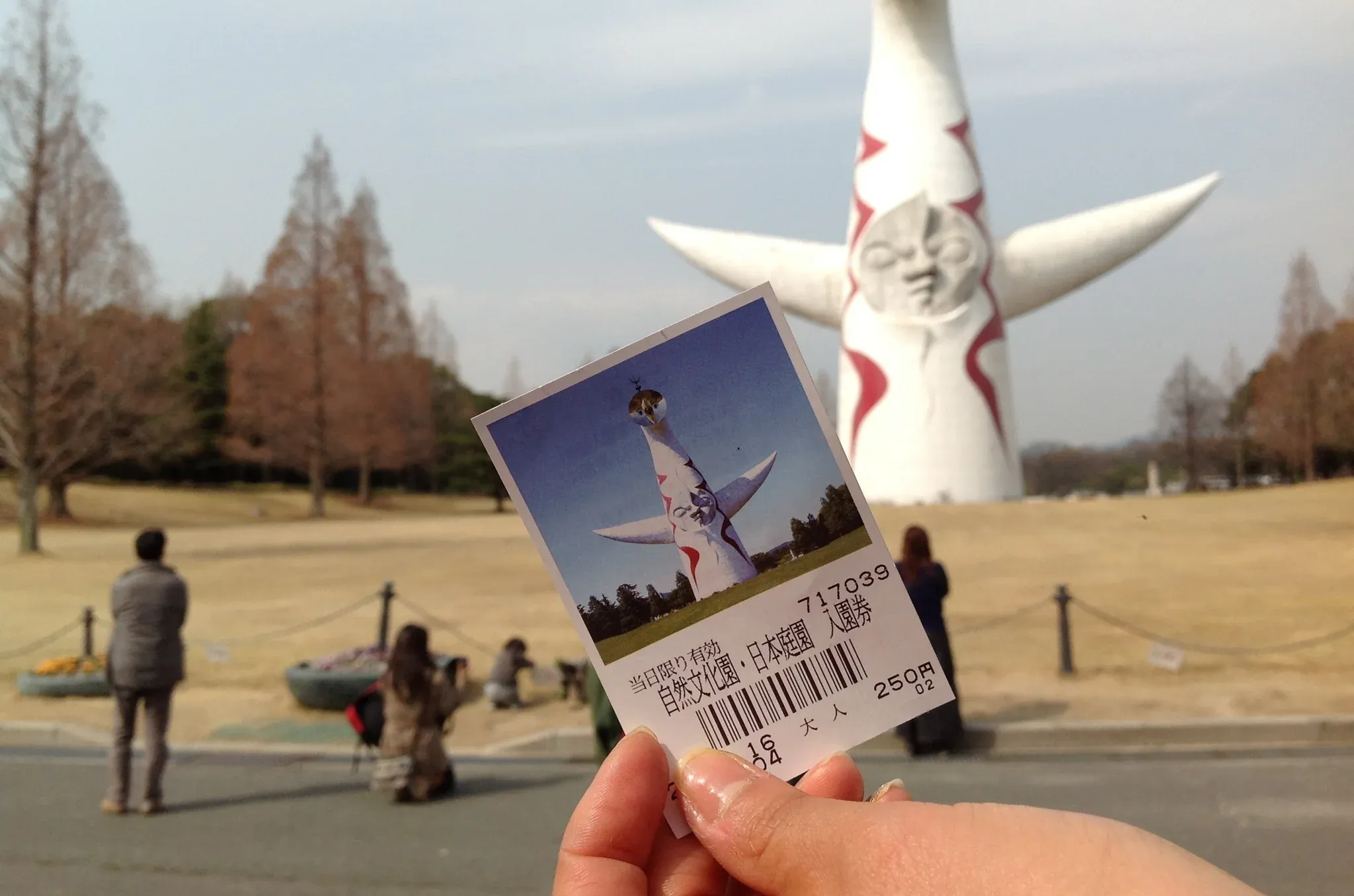 A hand holding an admission ticket to Expo ’70 Commemorative Park with the Tower of the Sun rising in the background.