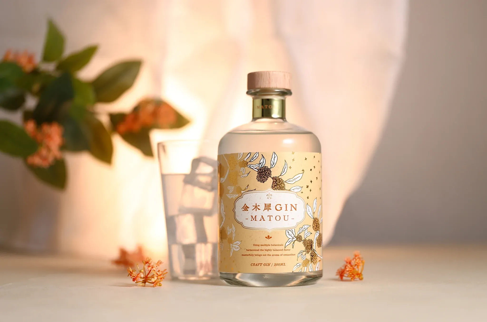 A bottle of Kinmokusei GIN -MATOU- stands beside a glass of ice water on a softly lit surface, surrounded by small orange osmanthus blossoms and green leaves in the background.