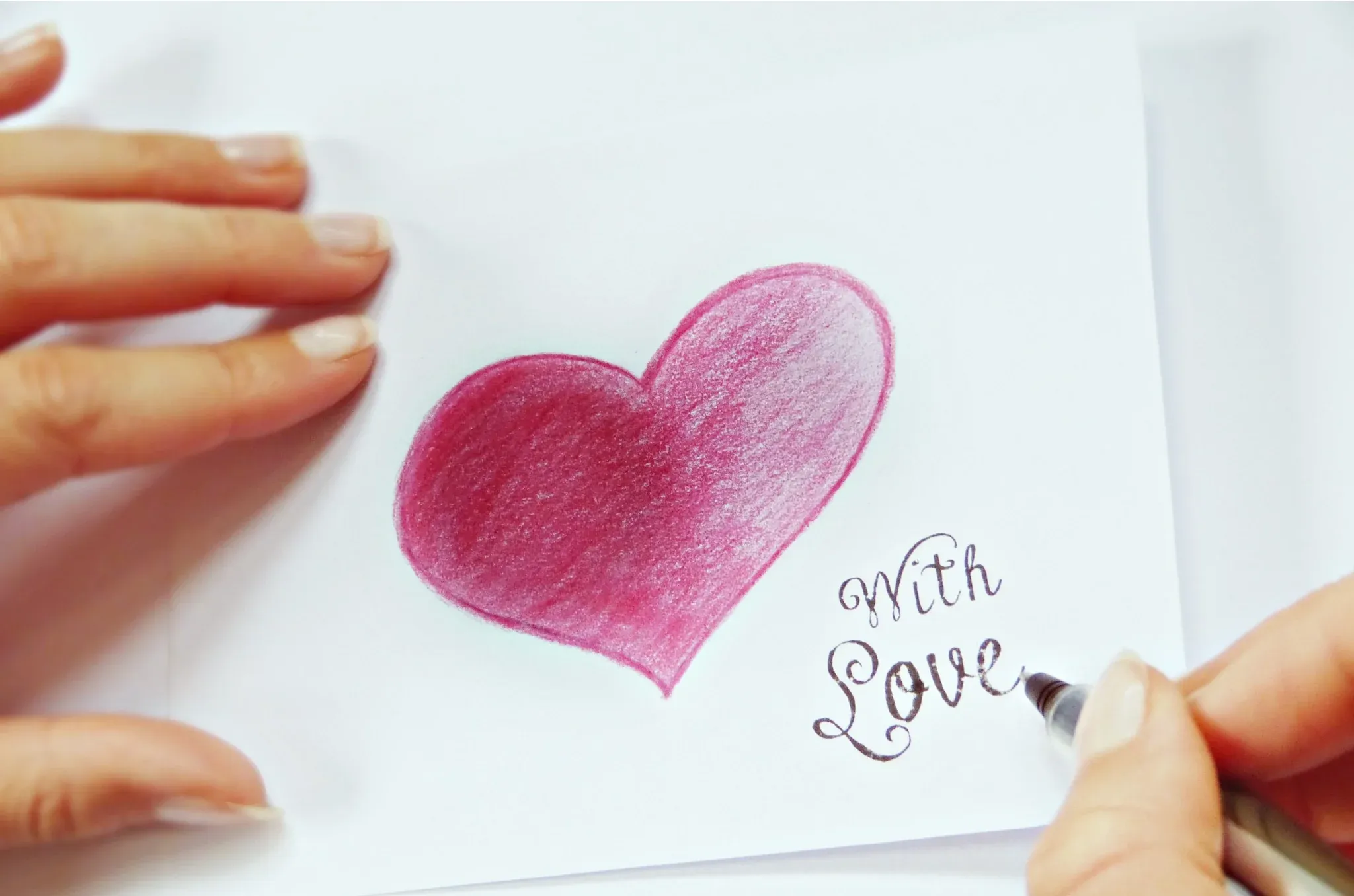 A hand drawing a pink heart and writing “with Love” on paper.