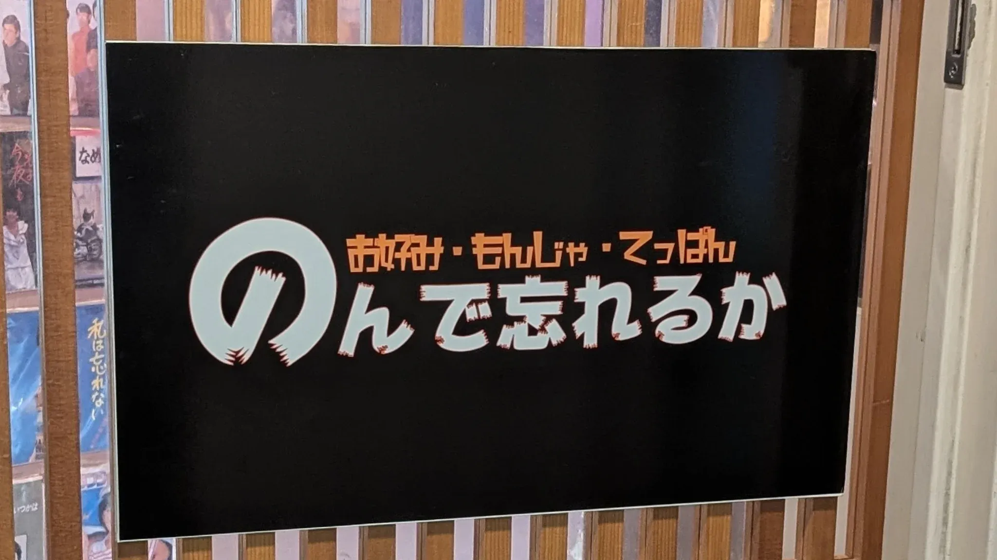 黒い看板に「お好み・もんじゃ・てっぱん のんで忘れるか」と書かれている。