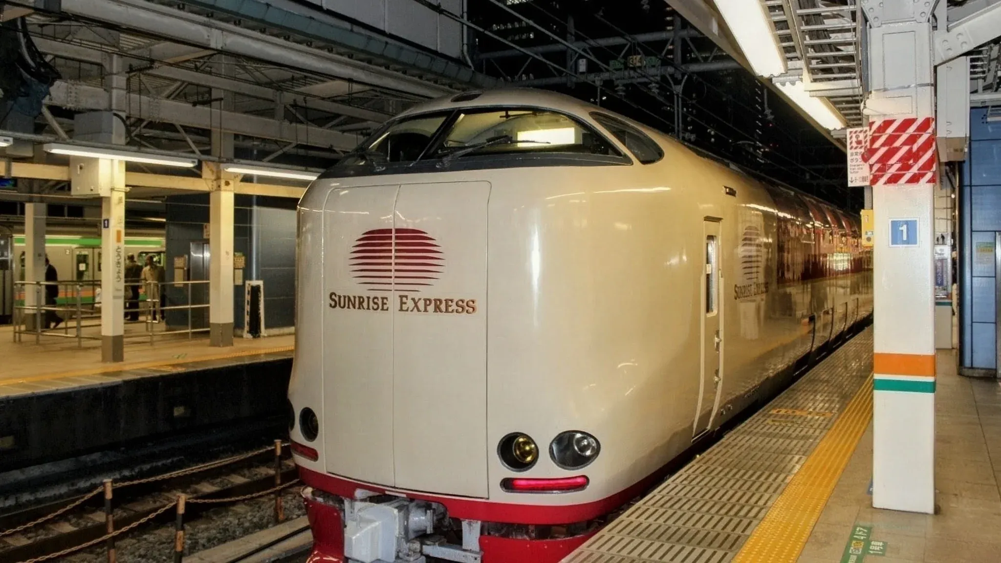 The front of the beige Sunset Express train at a station platform, featuring its rounded nose and red sun logo under bright lights.