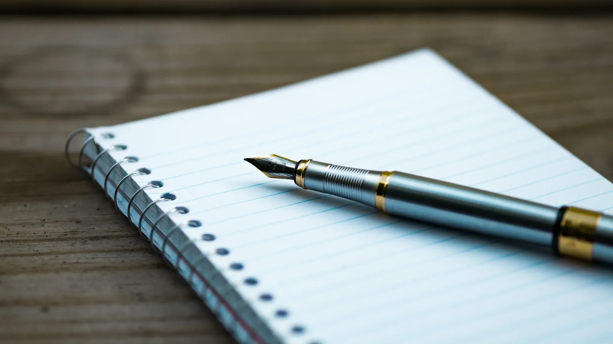A silver fountain pen resting diagonally on a blank spiral notebook placed on a wooden table.